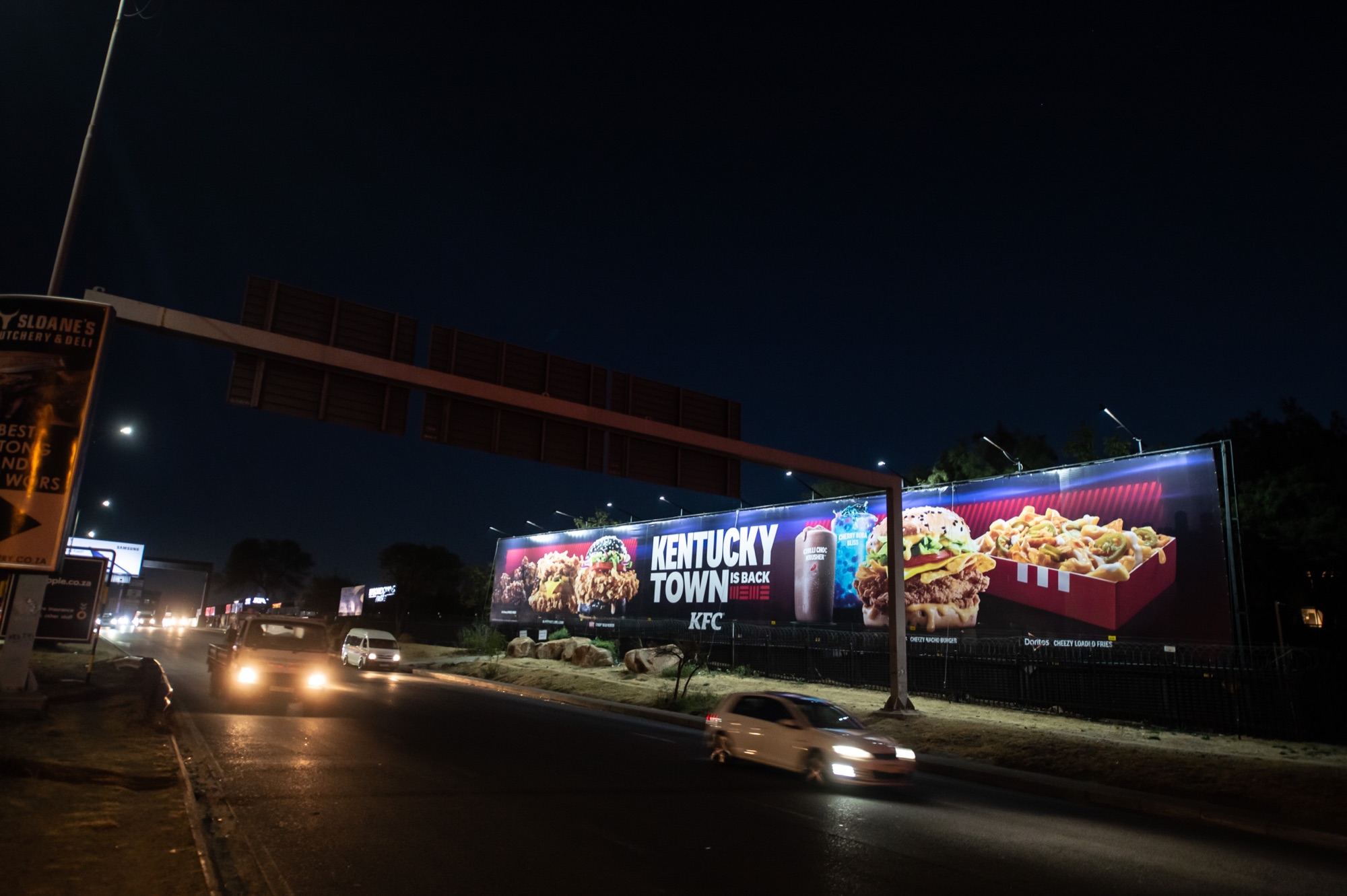 Illuminated KFC "Kentucky Town is Back" billboard on a New Wave Outdoor Media site at night in Johannesburg